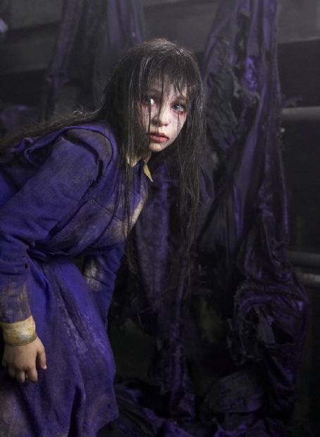 a woman with dark hair and makeup sitting on the ground in front of purple cloths