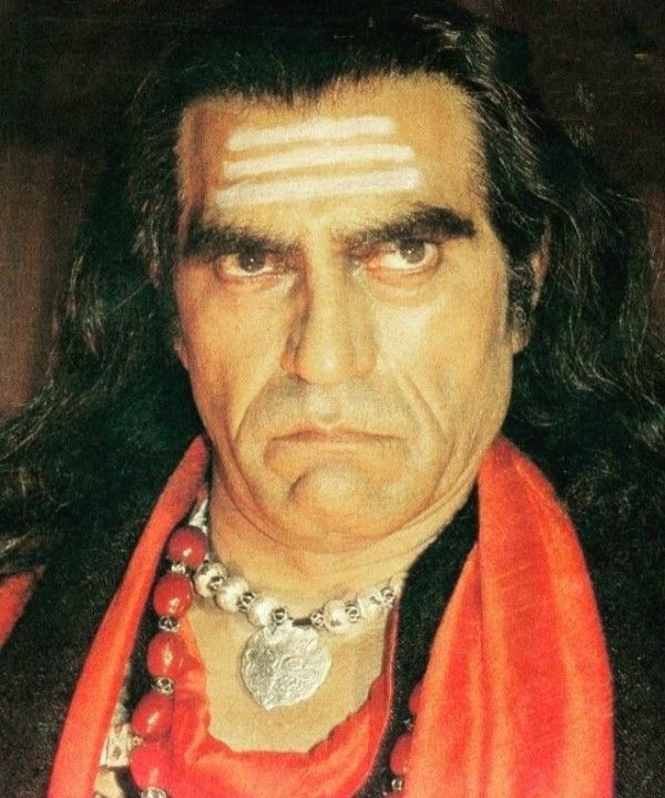 a man with long black hair wearing an orange scarf and beads around his neck is staring at the camera