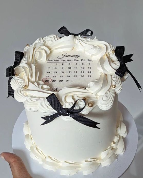 a white wedding cake with black ribbons and a calendar on the top is being held by a person's hand