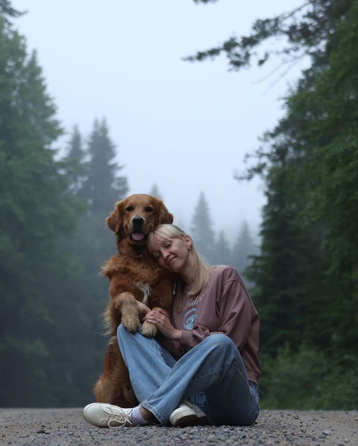 a woman sitting on the ground with her dog in front of trees and foggy sky