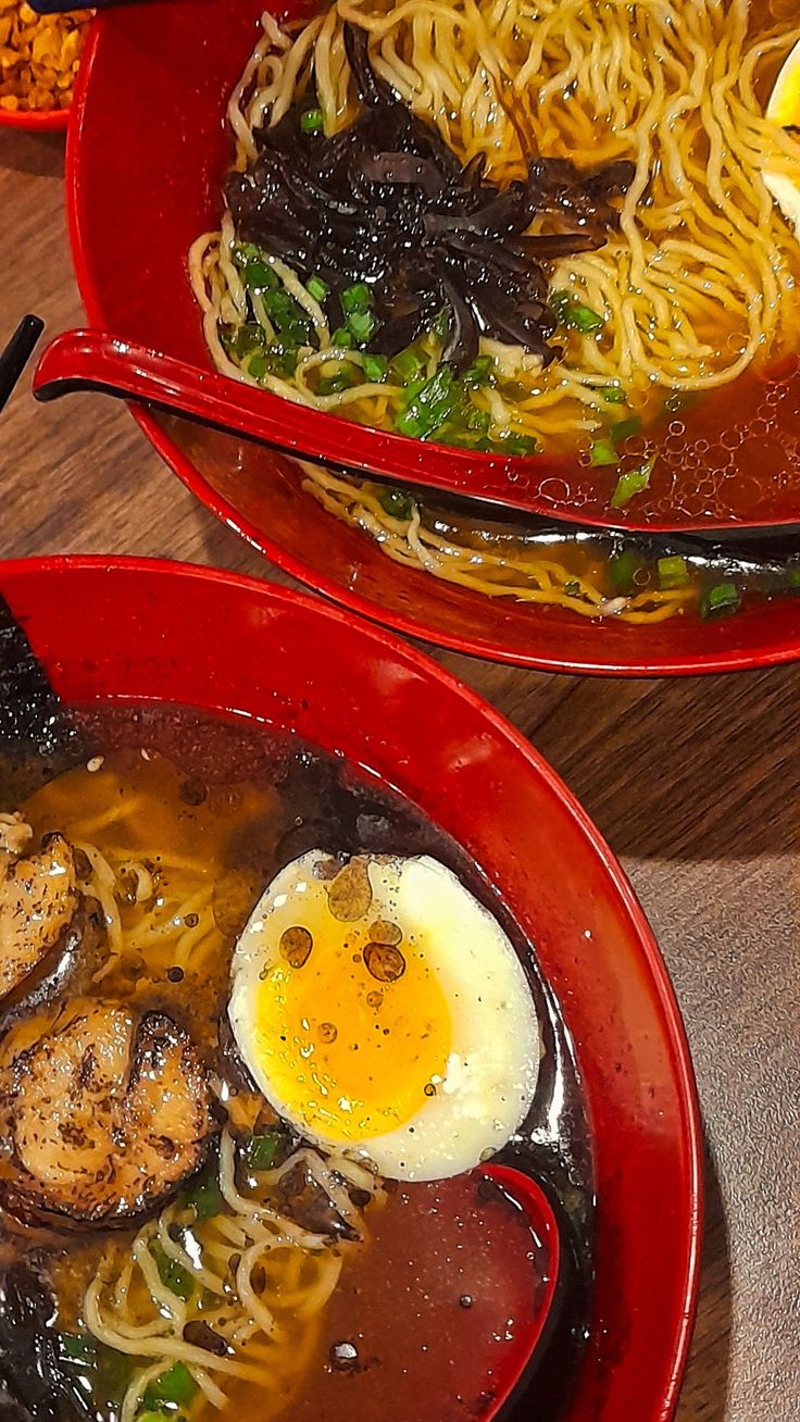 two red bowls filled with noodles, meat and an egg sitting on top of a wooden table