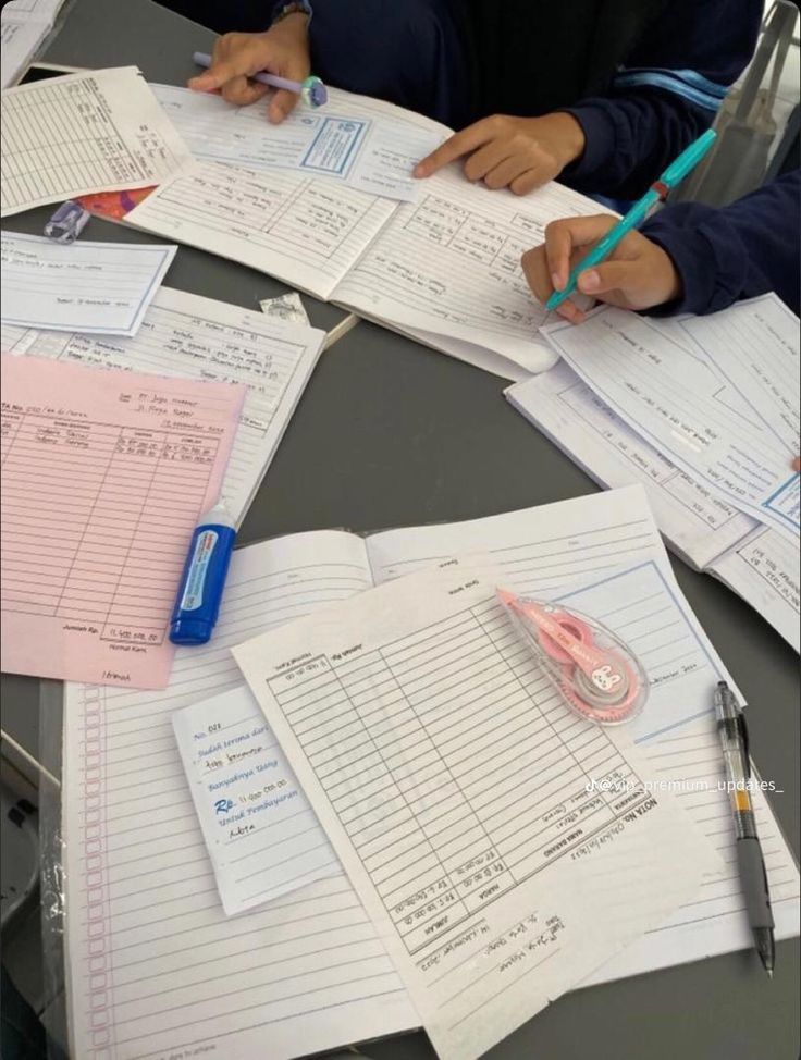 two people sitting at a table with papers and pens on top of each other,