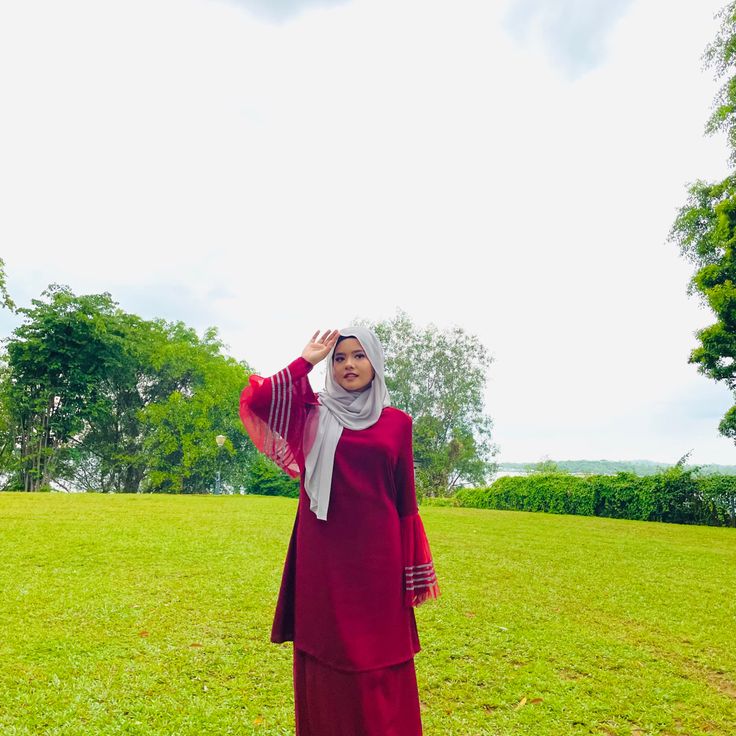 a woman in a red dress and head scarf standing on the grass with her hands behind her head