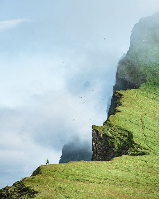 a man standing on top of a lush green hillside next to a tall mountain covered in fog