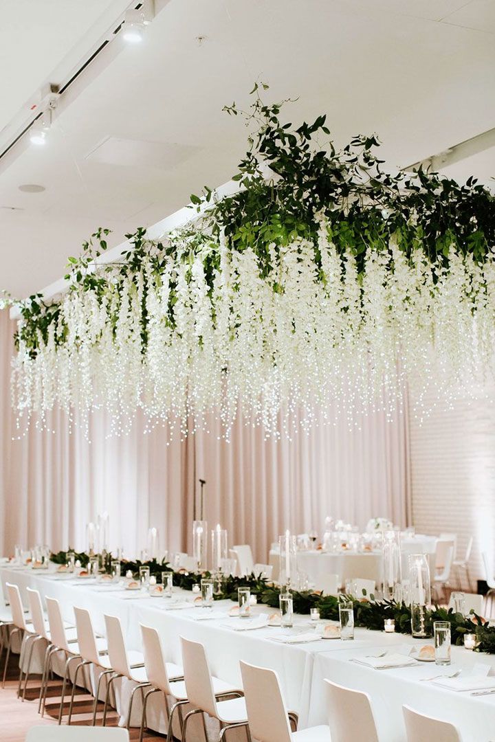 an elegant wedding reception with white flowers and greenery hanging from the ceiling, along with long tables