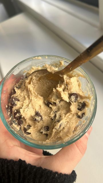 a hand holding a bowl with cookie dough and a wooden spoon in it, on top of a window sill