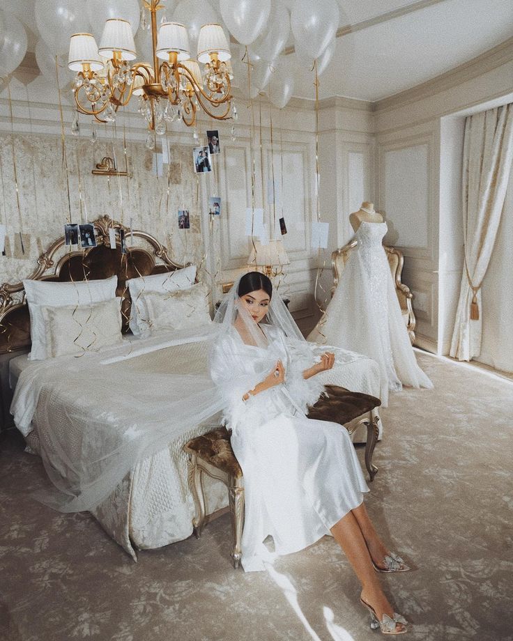 a woman sitting on top of a bed next to a wedding dress hanging from a chandelier