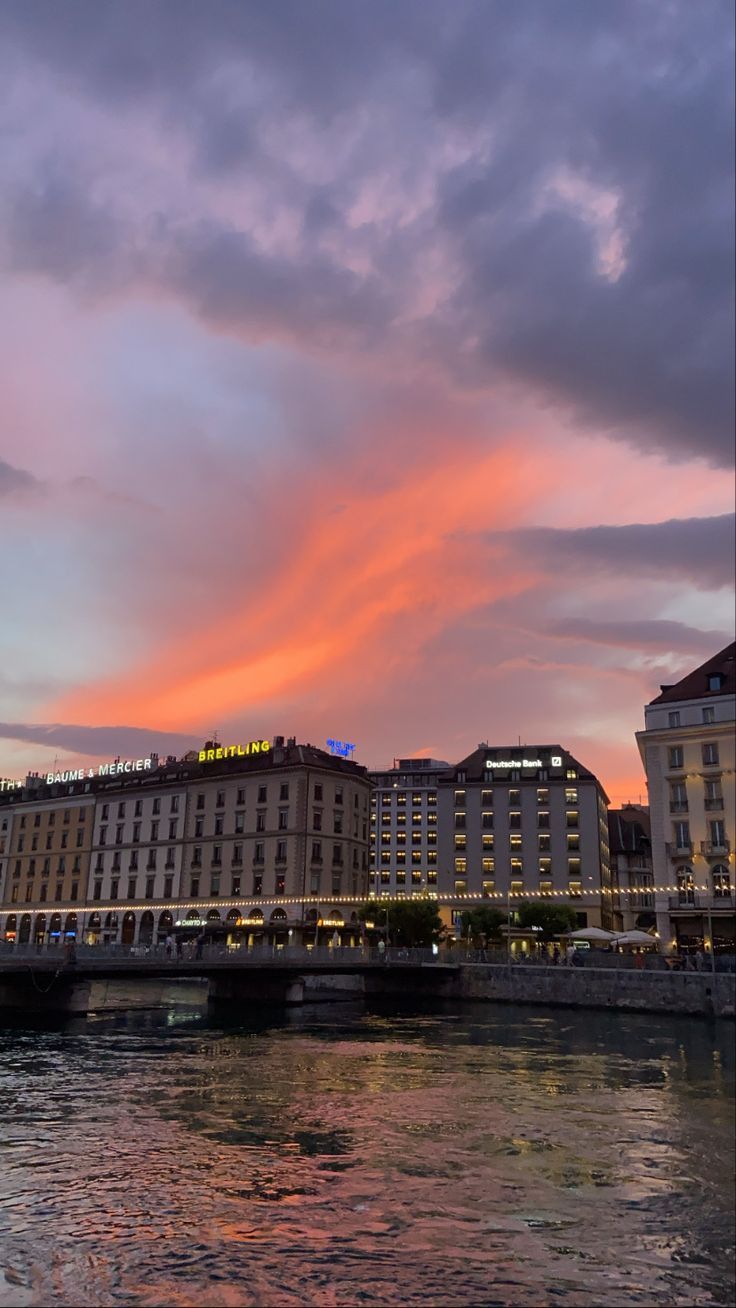 the sky is pink and purple as it sets over some buildings on the riverbank