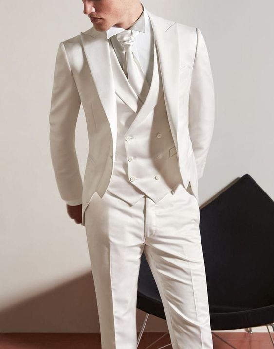 a man in a white tuxedo standing next to a black chair with his hands on his hips