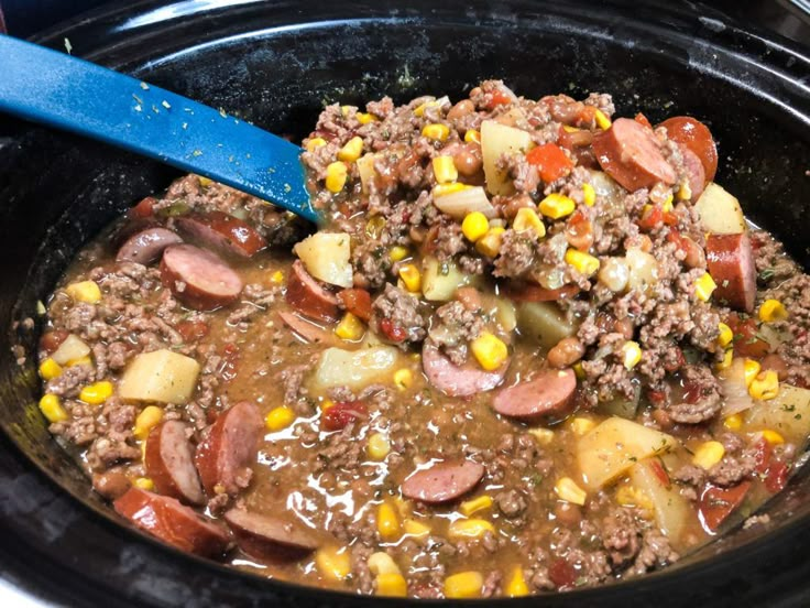 a crock pot filled with meat, potatoes and corn next to a blue spoon