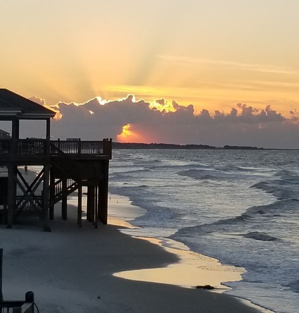 the sun is setting over the water at the beach with a pier in the foreground