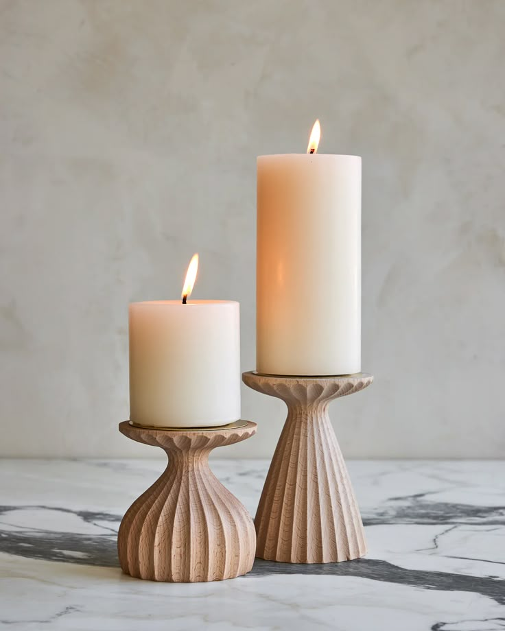 two white candles sitting next to each other on a marble counter top, with one candle lit in the middle