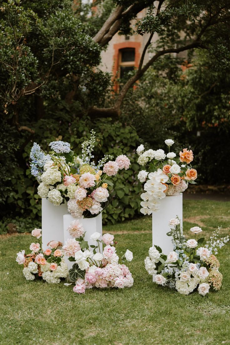 three white vases with flowers are on the grass in front of some bushes and trees