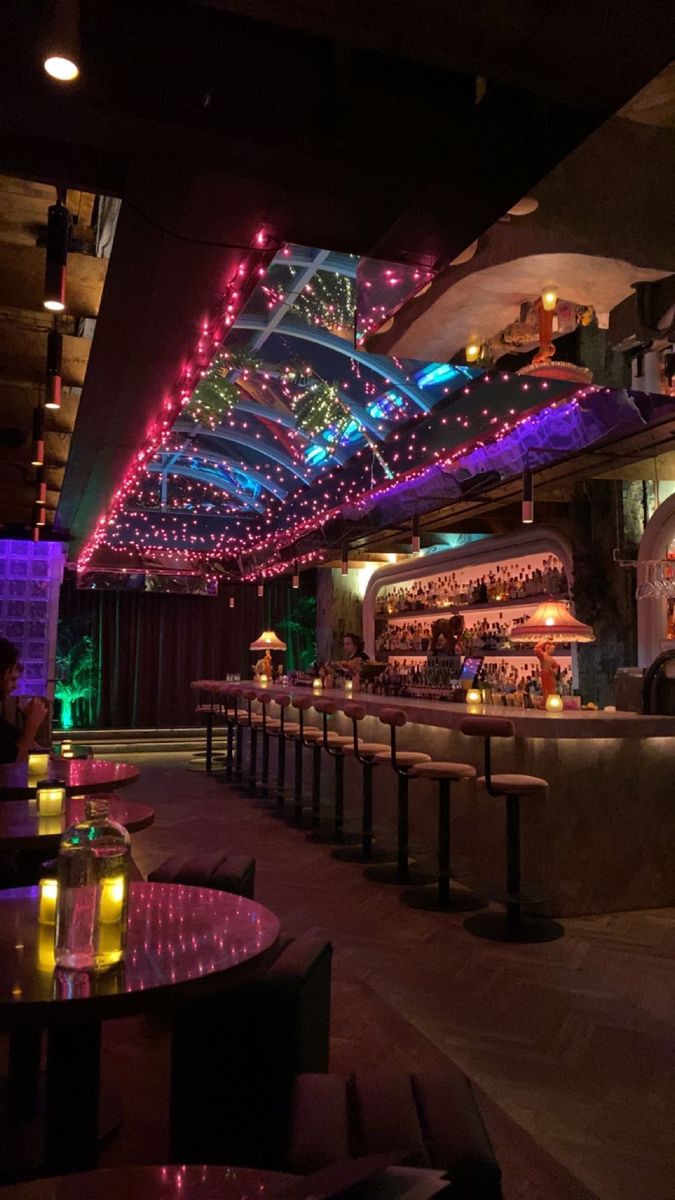 a dimly lit bar with colorful lights on the ceiling and tables in front of it