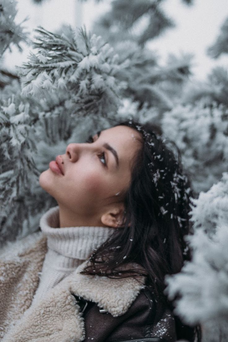 a woman standing in the snow next to a tree with her eyes closed and she is looking up