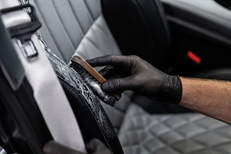 a person in black gloves cleaning the inside of a car seat with a brush and cloth