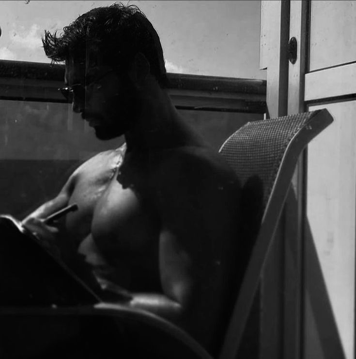 a shirtless man sitting in a chair writing on a piece of paper next to a window