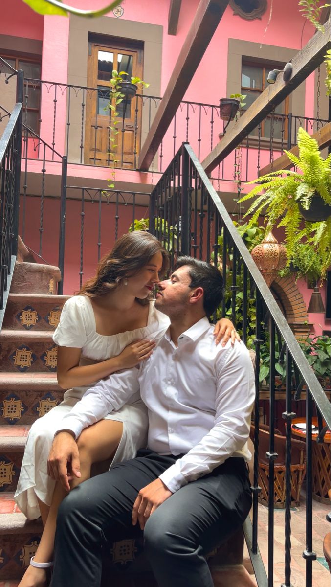 a man and woman are sitting on the steps in front of a pink building, kissing