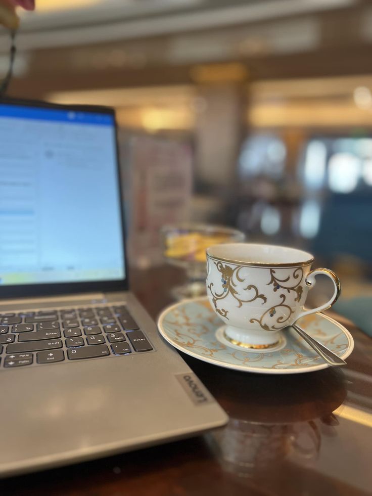 a laptop computer sitting on top of a wooden table next to a cup and saucer