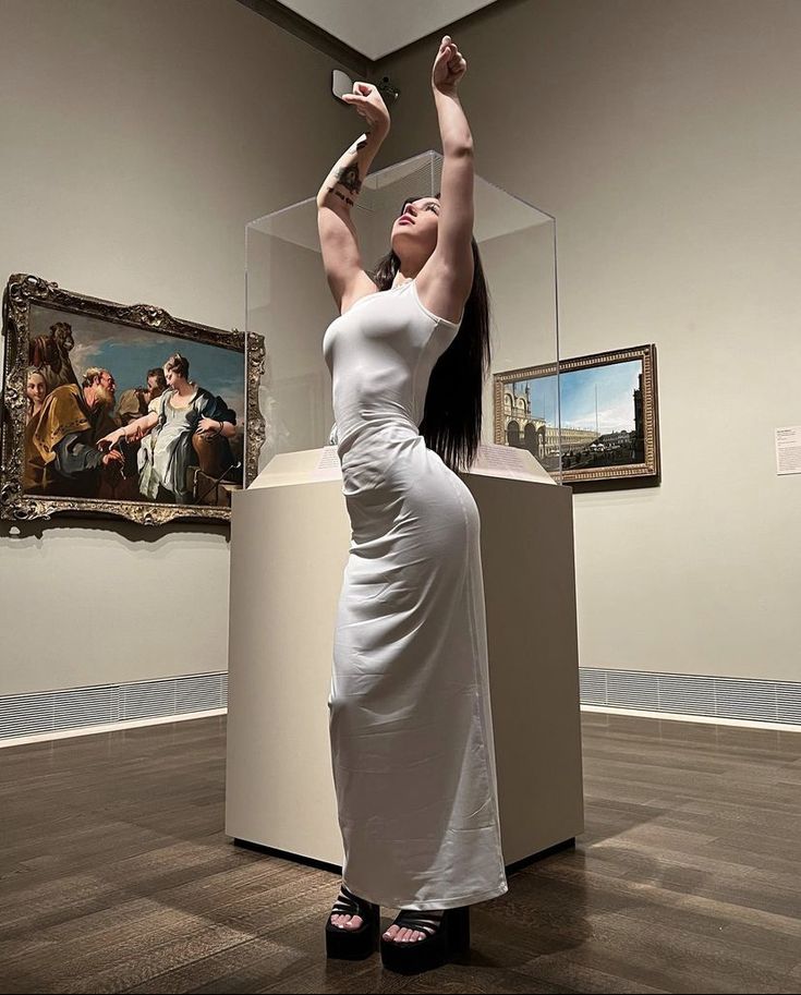 a woman in a white dress is holding her arms up while looking at some paintings