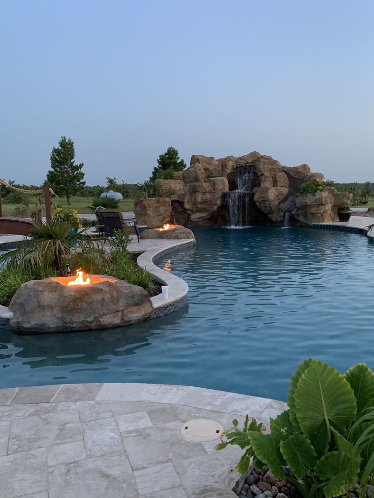 a pool with a waterfall and fire pit in the middle, surrounded by greenery