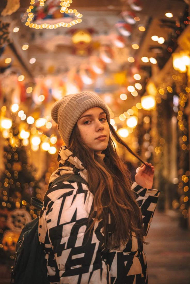 a woman standing in front of christmas lights wearing a beanie and looking at the camera