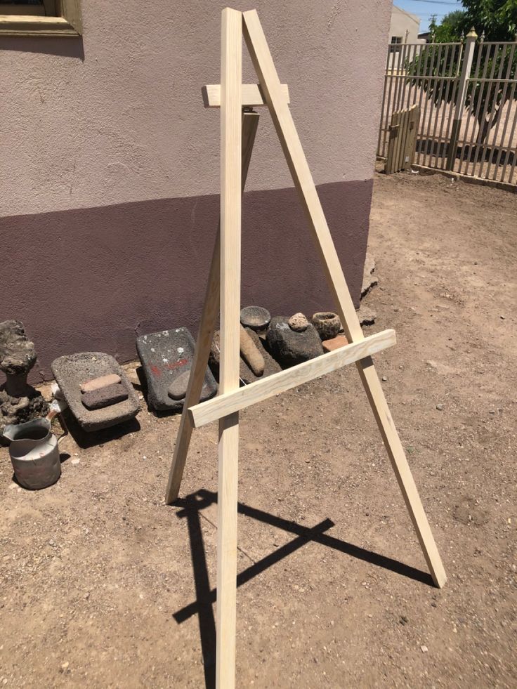 a wooden easel sitting on top of a dirt field next to a building and potted plants