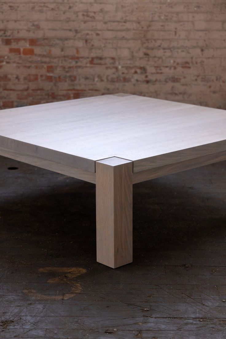 a square wooden table sitting on top of a floor next to a brick wall in an empty room