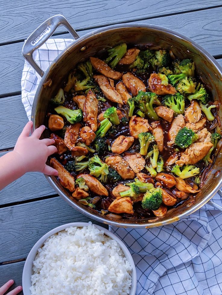 a pan filled with chicken, broccoli and rice next to a child's hand