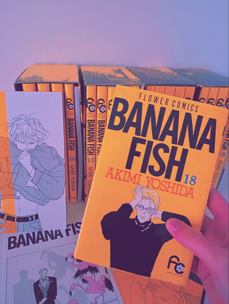 a person holding up a banana fish book in front of several other books on the table