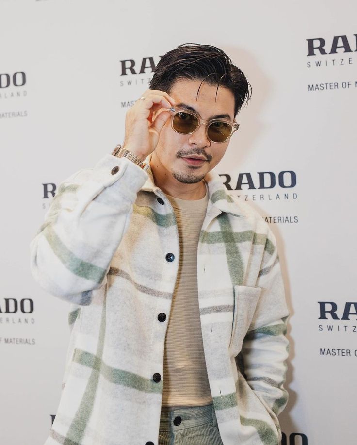 a man with sunglasses on standing in front of a white wall holding his hand up to his ear