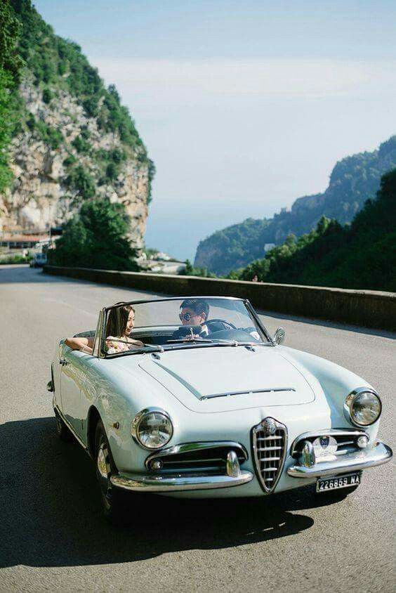 a man and woman driving in an old convertible car on the road with mountains in the background