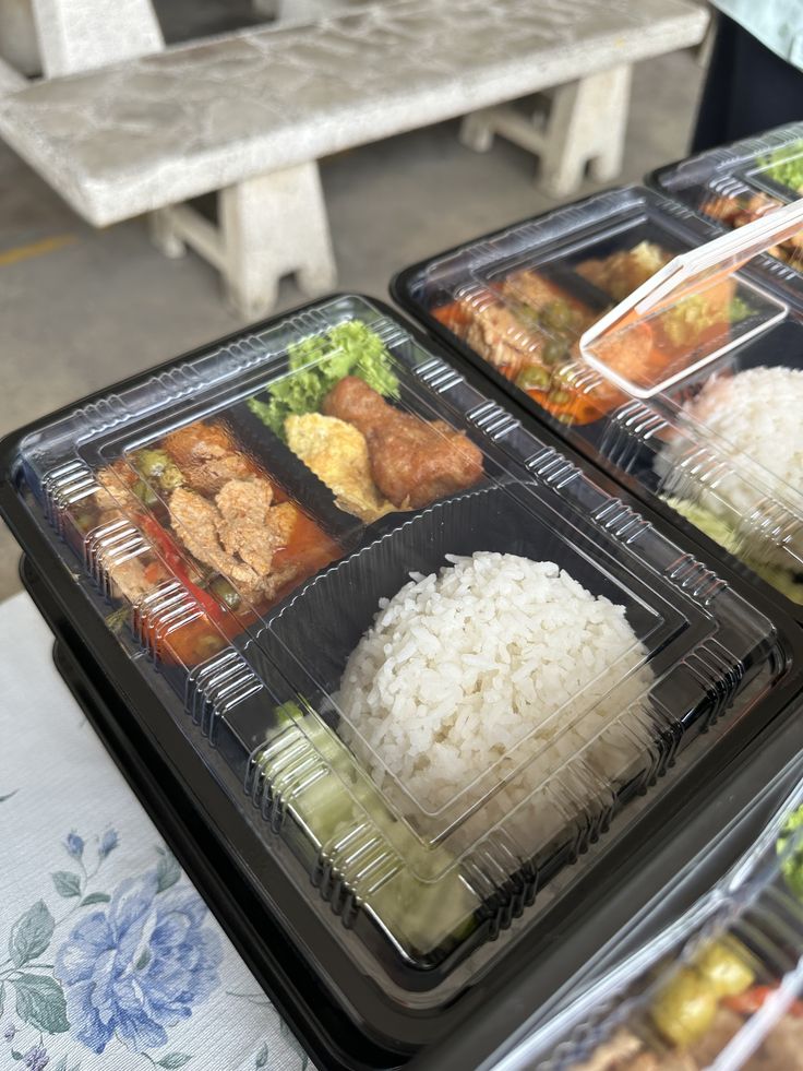 two plastic trays filled with rice and meat on top of a table next to a bench