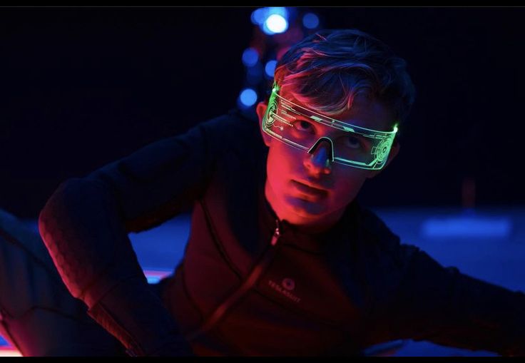 a young man wearing glowing glasses in the dark with his hands on his hips looking at something