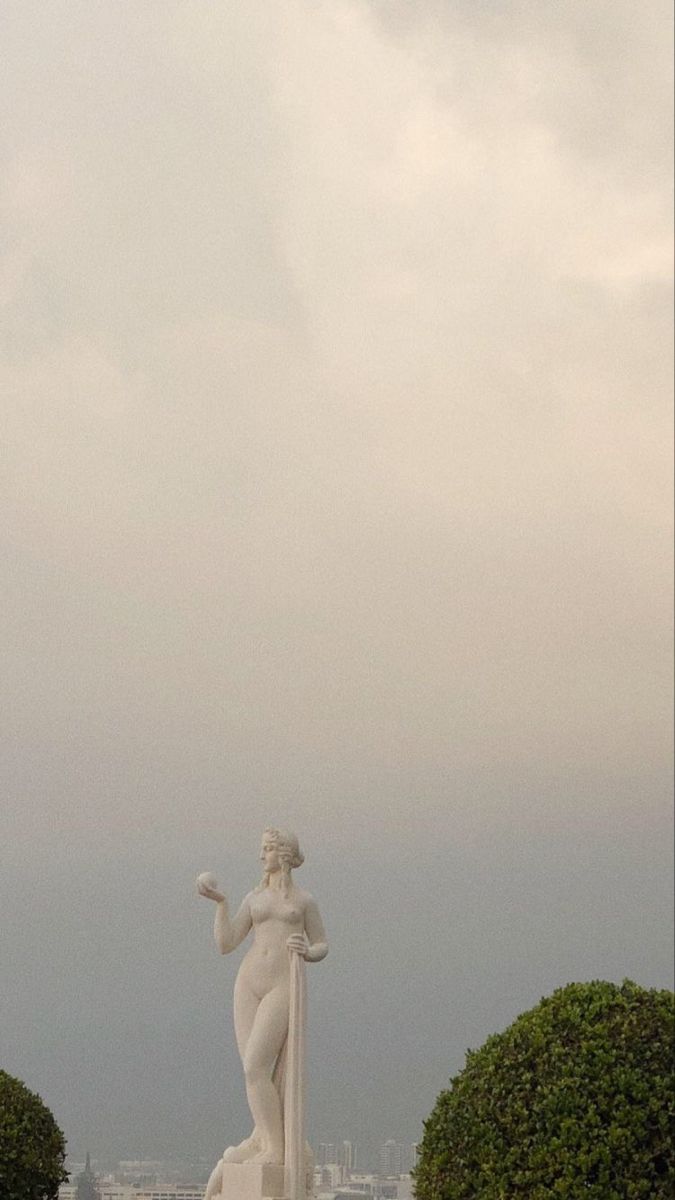 there is a white statue in the middle of some bushes and trees with buildings in the background