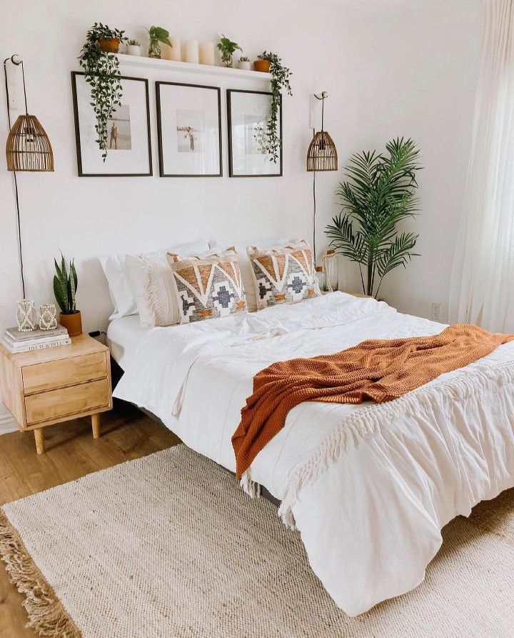 a bed with white sheets and pillows in a bedroom next to a wooden dresser, potted plants and pictures on the wall