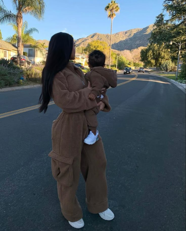 a woman holding a baby in her arms on the street with palm trees and mountains in the background