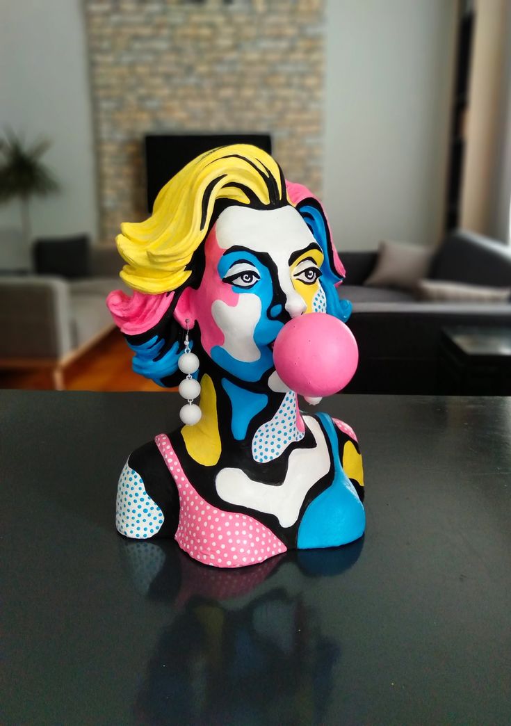 a statue of a woman blowing a bubble with her face painted blue and yellow, sitting on a table in front of a fireplace