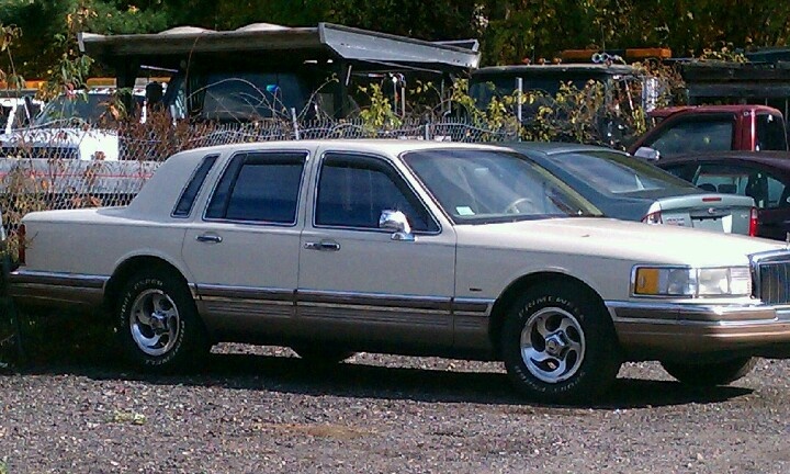 a white car parked in a parking lot next to some trees and bushes with other cars behind it