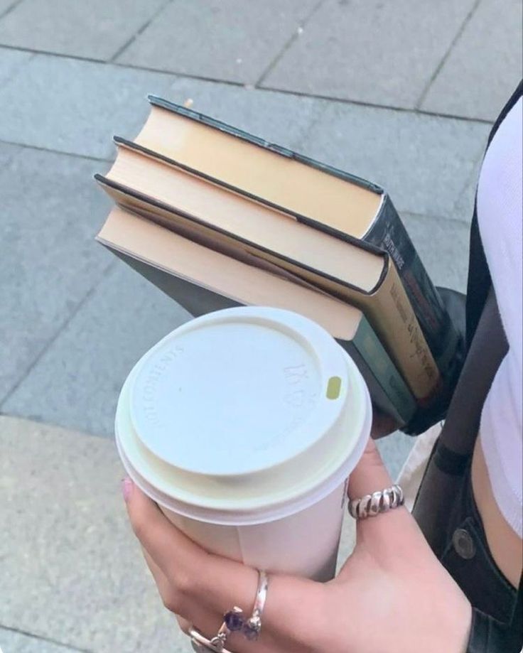 a woman holding a cup of coffee and several books on her arm while walking down the street