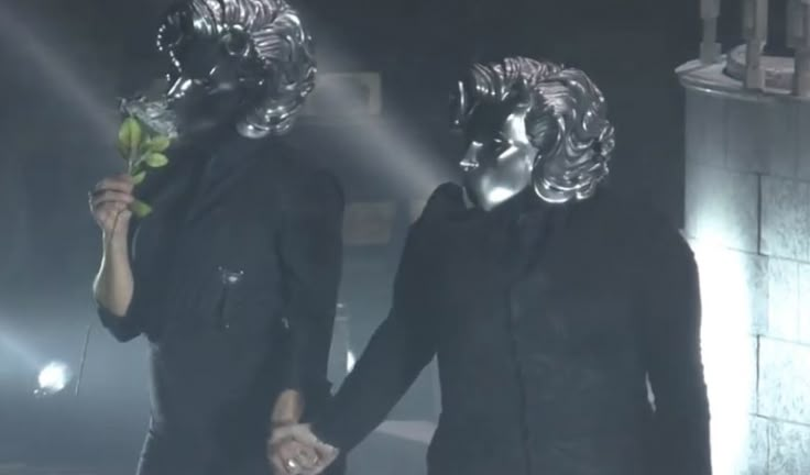 two people with silver masks holding hands in front of a light and some lights behind them