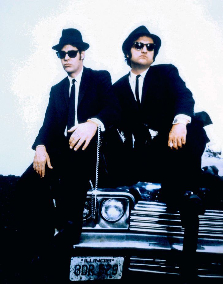 two men in suits and hats sitting on the hood of a car, one with his arm around another man's shoulder