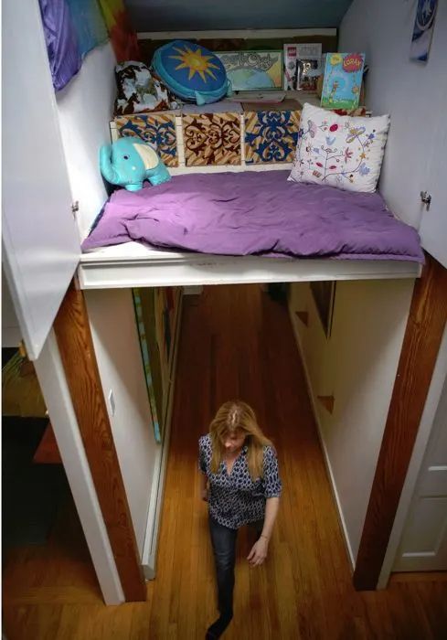 a woman standing under a bed in a room with wooden floors and purple bedspread