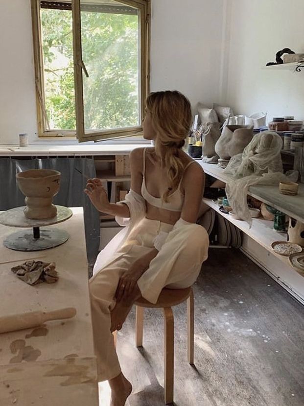 a woman sitting on a chair in a room with pottery and other items around her