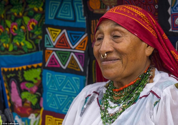 an older woman wearing a red headdress and nose ring in front of colorful wall hangings