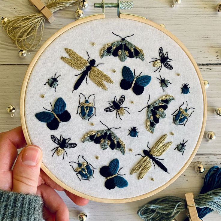 a person holding up a cross - stitch project with bugs and moths on it, surrounded by other crafting supplies