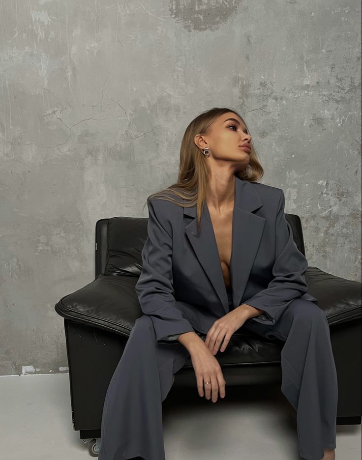 a woman sitting on top of a black couch in front of a cement wall wearing a suit