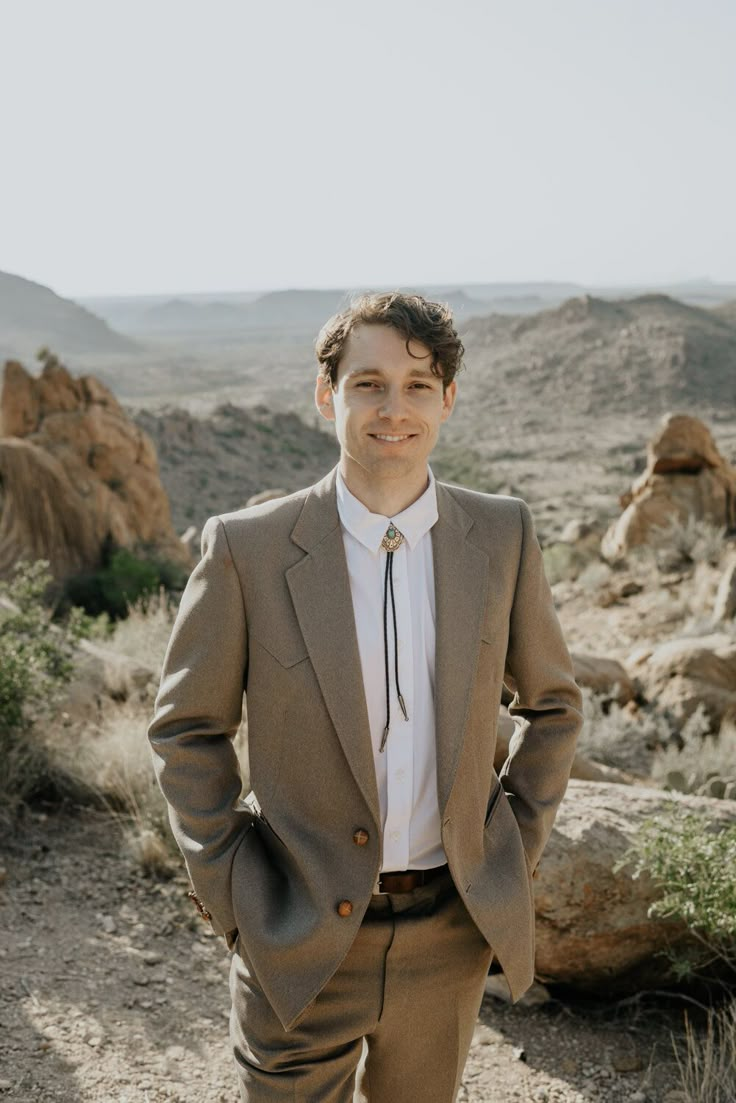 a man standing in the desert with his hands in his pockets, wearing a suit and tie