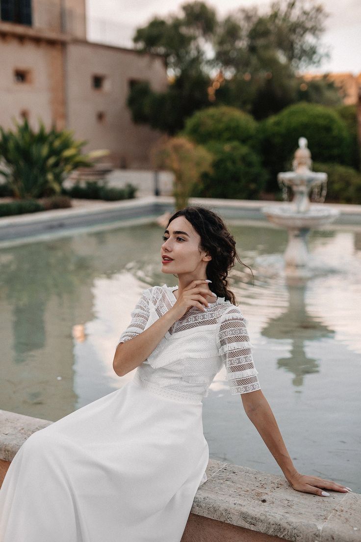 a woman in a white dress leaning against a wall next to a water fountain and looking off into the distance