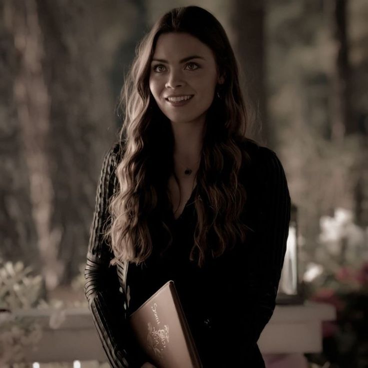 a woman with long hair holding a folder and smiling at the camera while standing in front of a tree